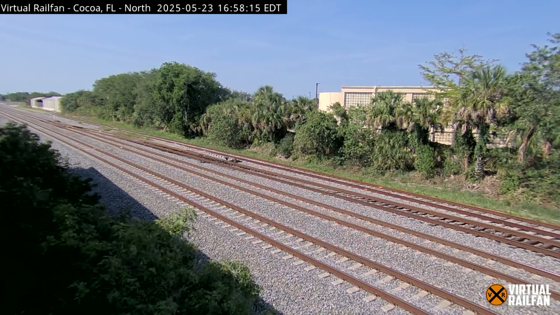 Multiple train tracks stretch into the distance under a clear sky, passing by lush green vegetation and a building.