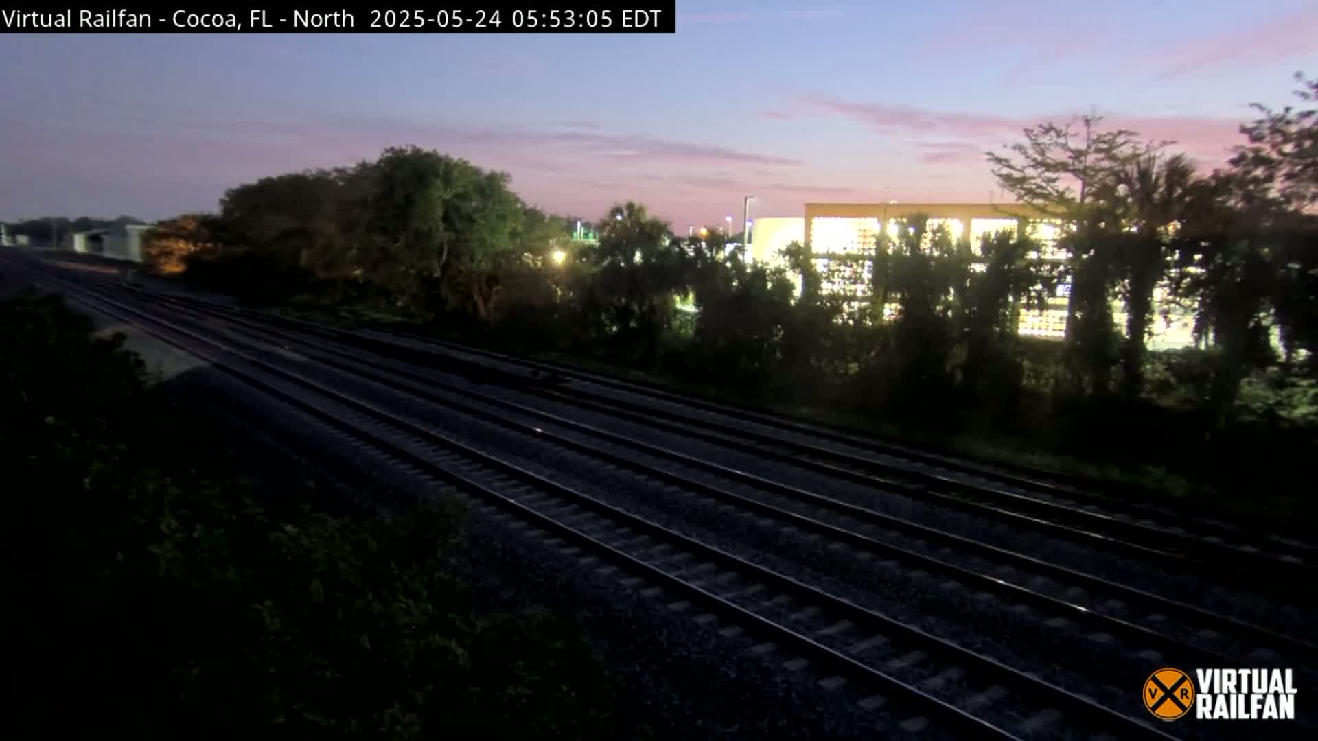 Multiple train tracks extend into the distance under a twilight sky, bordered by trees and a large building.