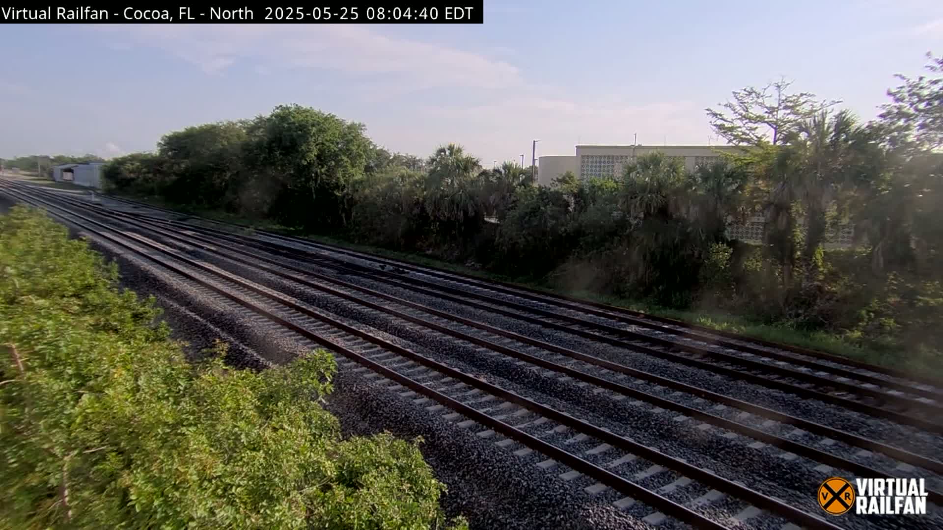 Multiple train tracks stretch into the distance under a partly sunny sky, bordered by lush green vegetation and a distant building.