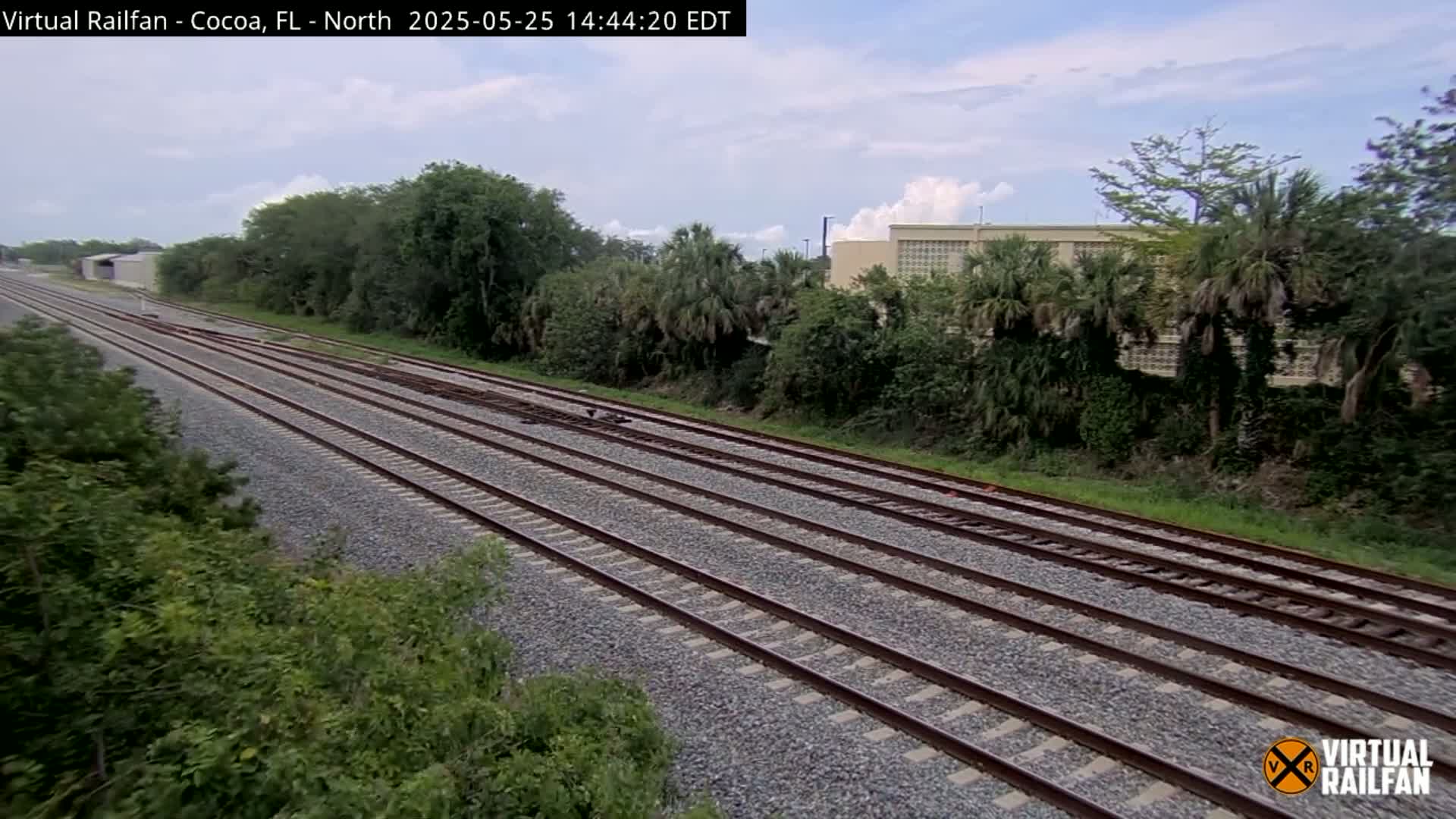 Multiple parallel train tracks stretch into the distance under a partly cloudy sky, bordered by lush green vegetation and a building in the background.