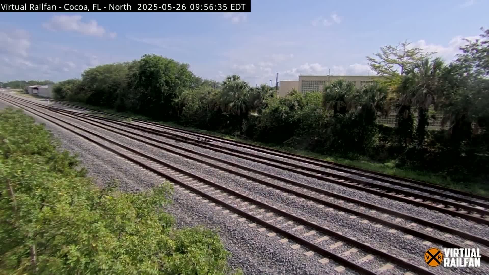 Multiple train tracks extend into the distance under a partly cloudy sky, bordered by lush green vegetation and a building in the background.