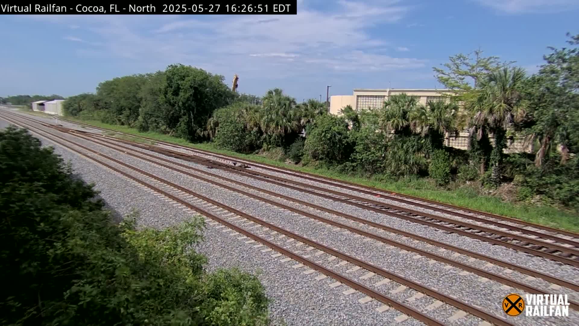 Multiple train tracks stretch into the distance under a partly sunny sky, flanked by lush green vegetation and a building in the background.