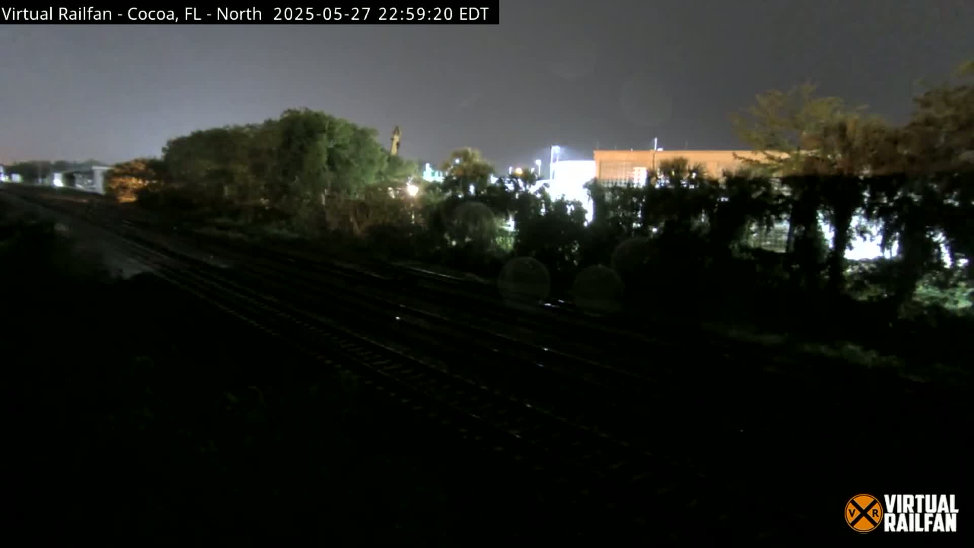 A nighttime view of railroad tracks running alongside dark vegetation, with a dimly lit building visible in the background under a dark sky.