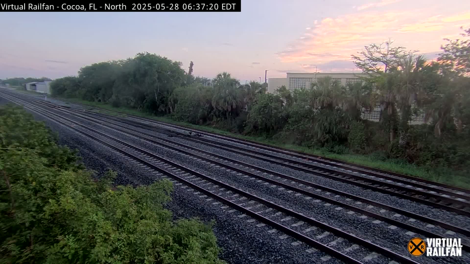 Multiple train tracks extend into the distance under a sunrise sky, bordered by lush green vegetation and a building in the background.