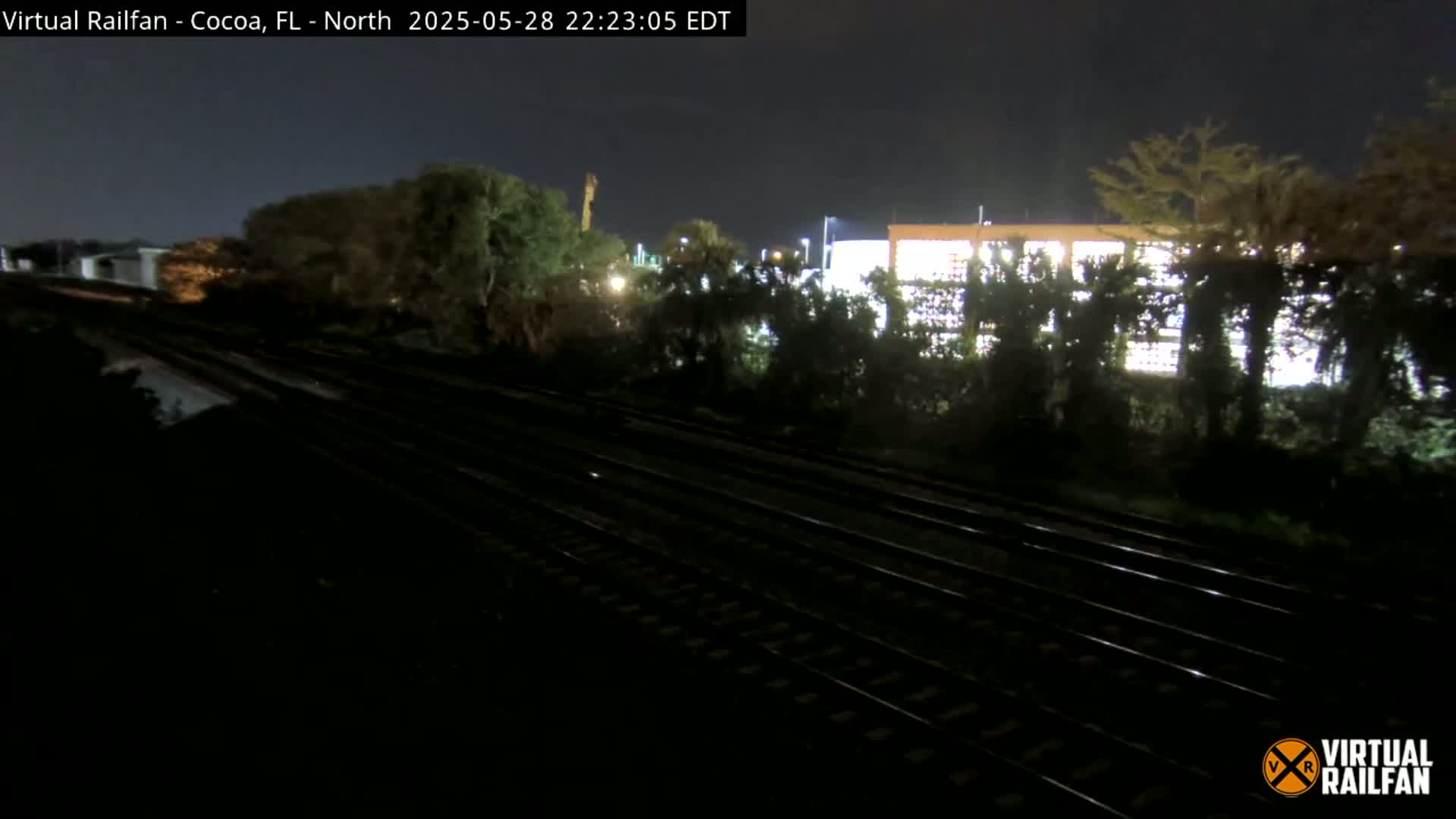At night, a view of multiple train tracks runs horizontally through the center of the image, with dark vegetation and a brightly lit building visible in the background.