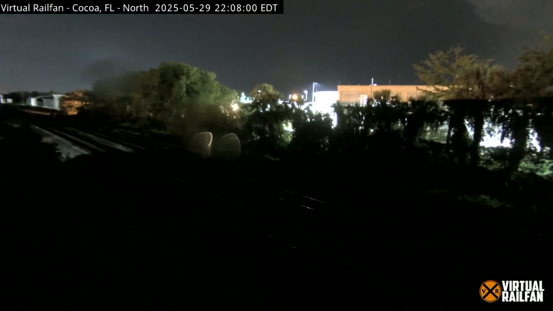 A nighttime view of railroad tracks running alongside dark trees and a dimly lit building under a cloudy sky.