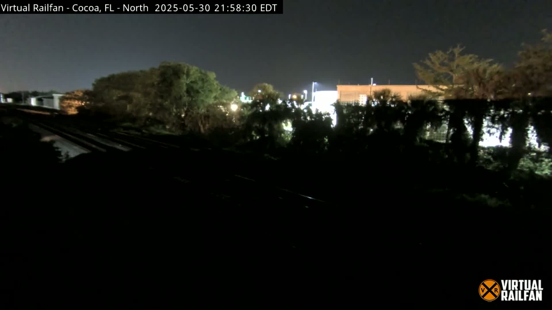 A nighttime view of railroad tracks bordered by dark, lush vegetation and dimly lit buildings in the distance under a clear sky.
