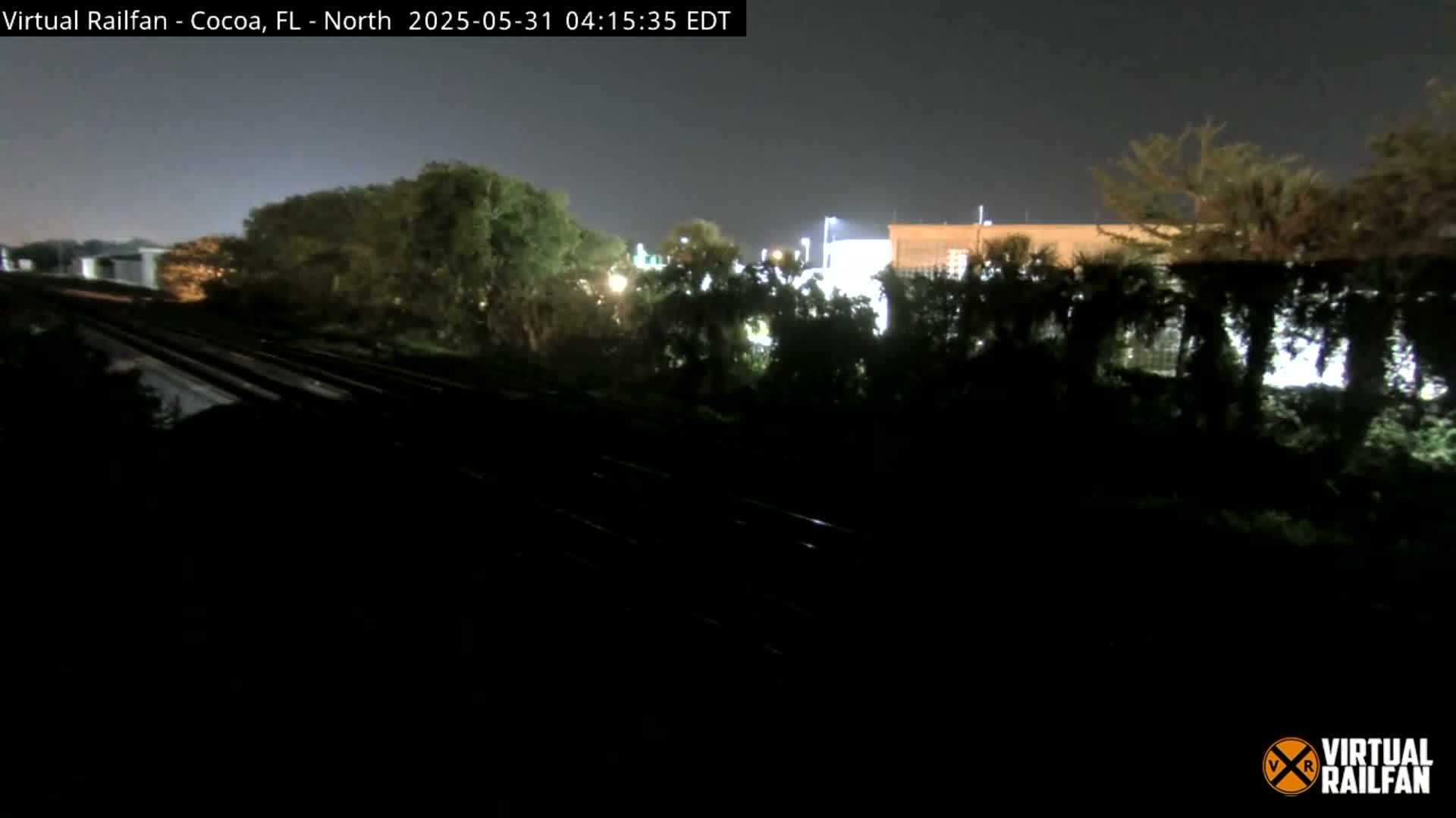 At night, a nighttime view shows train tracks running beside a line of trees and buildings under a dark sky.