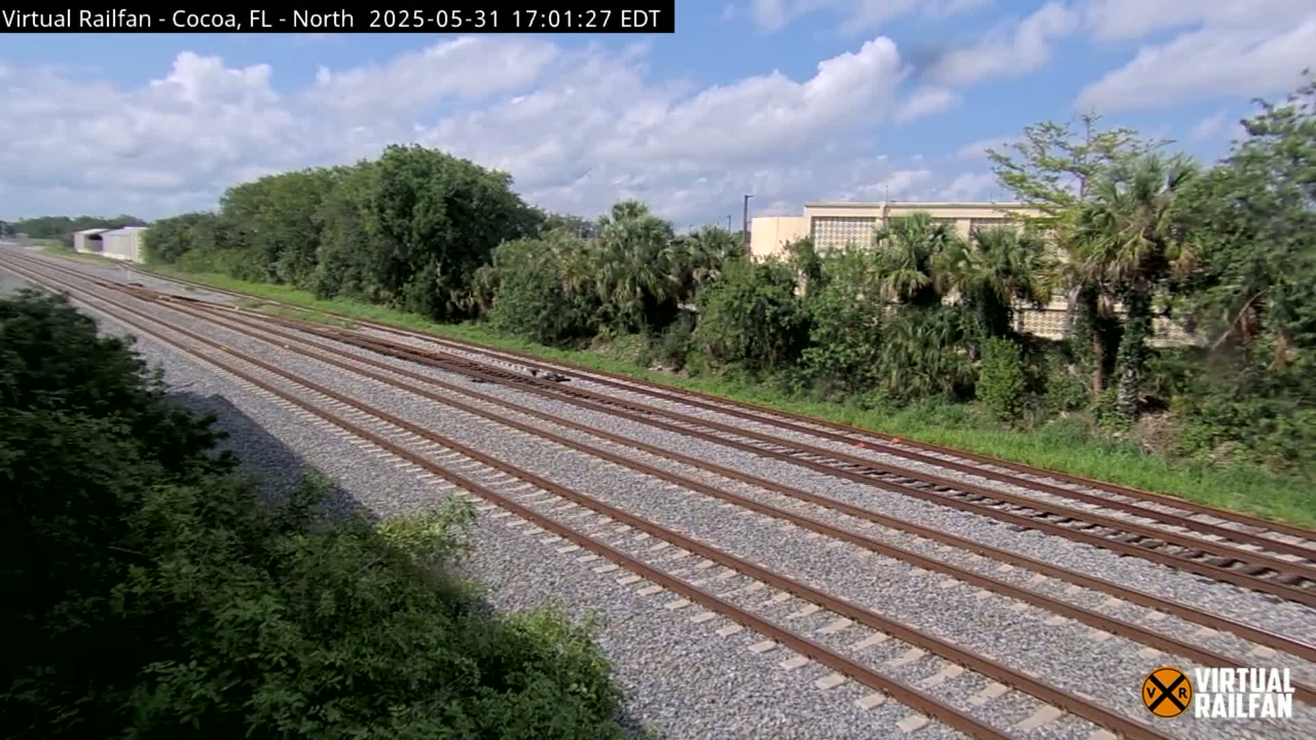 Multiple train tracks stretch into the distance, bordered by lush green vegetation under a partly cloudy sky.