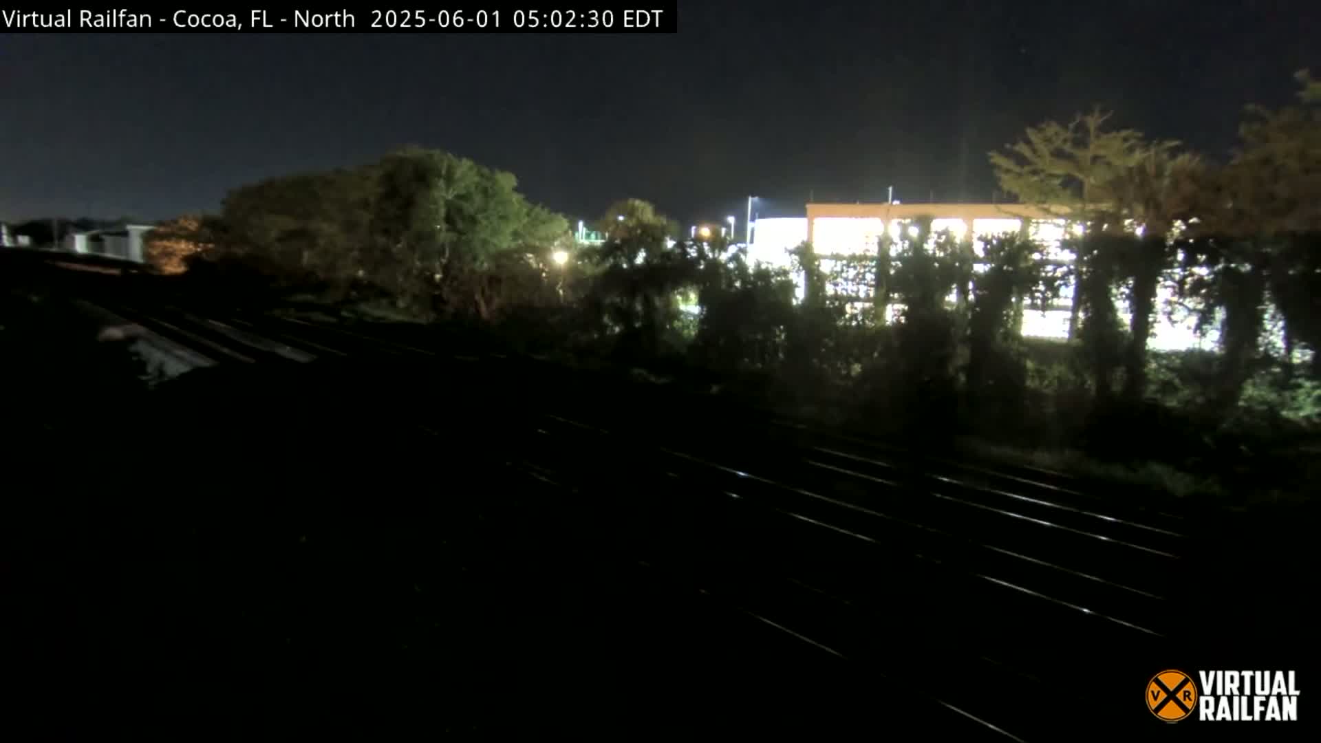 A nighttime view of railroad tracks beside a line of trees and a lit building under a clear, dark sky.