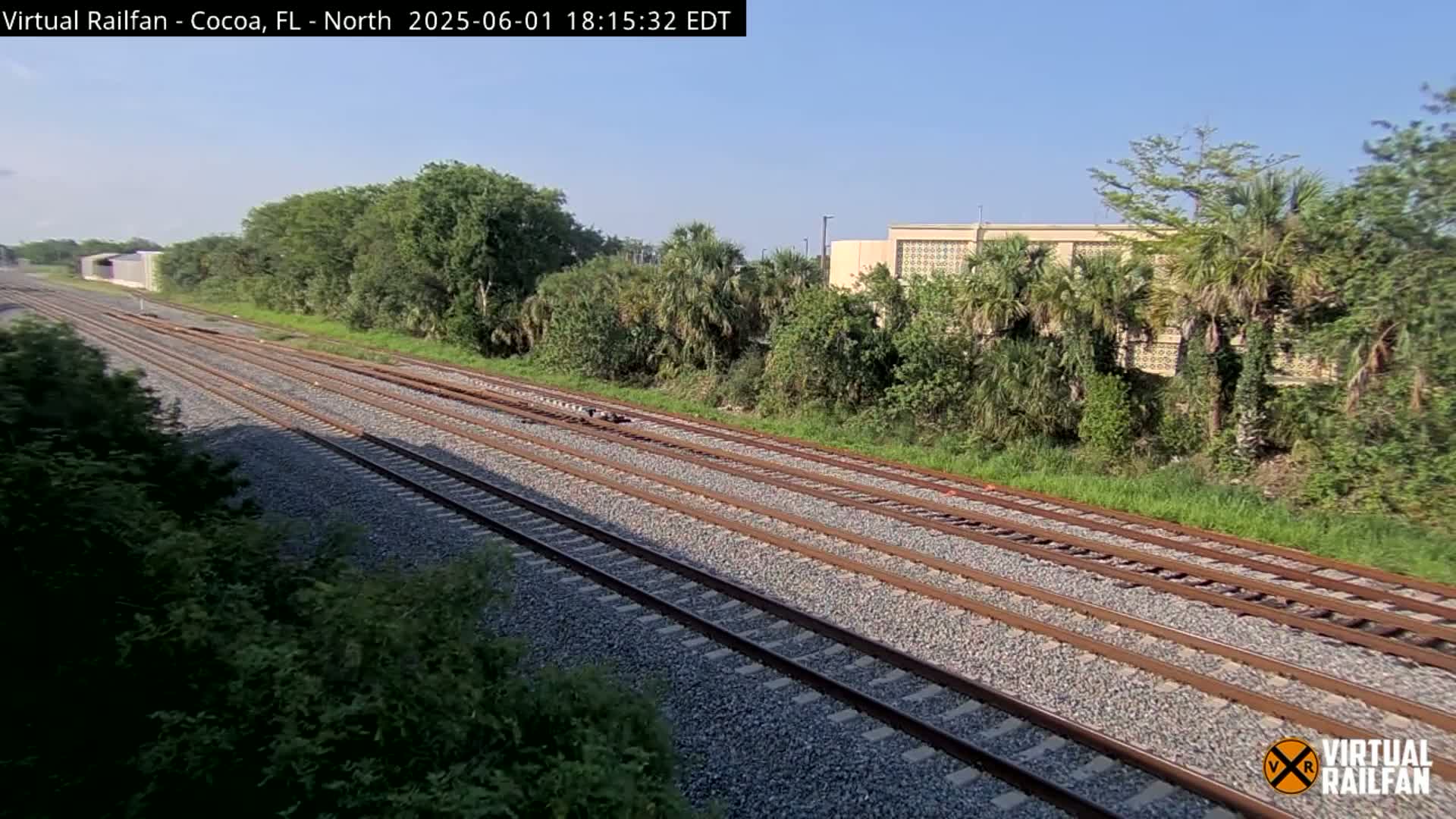 Multiple train tracks extend into the distance, bordered by lush green vegetation and a building under a clear sky.