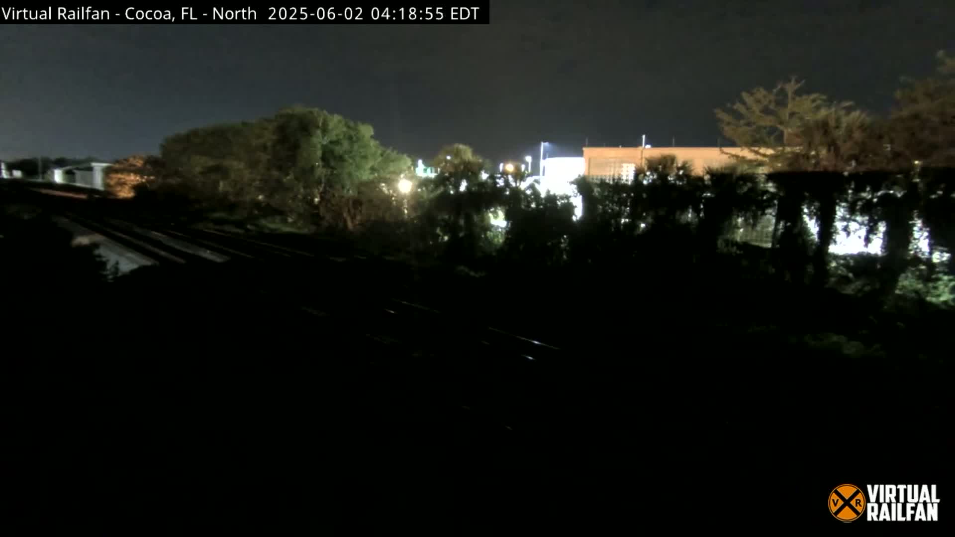 A nighttime view of railroad tracks running through a dark, tree-lined area near a building, under a clear sky.
