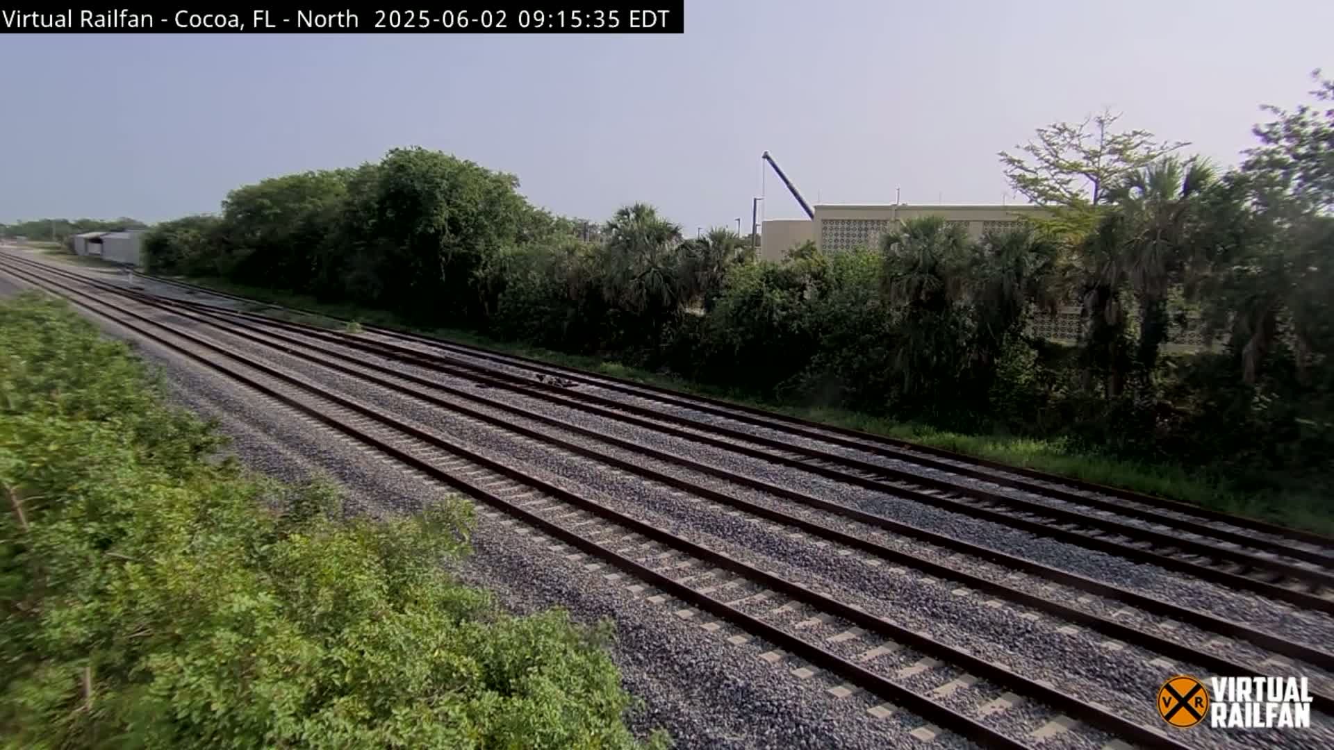 Multiple sets of train tracks stretch into the distance on a sunny day, bordered by lush green vegetation and a building in the background.