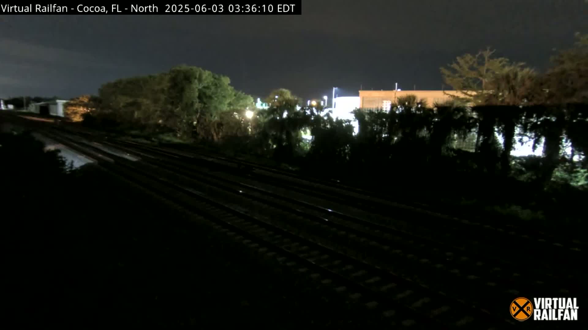 A nighttime view of multiple train tracks running through a dark, tree-lined area near a building.