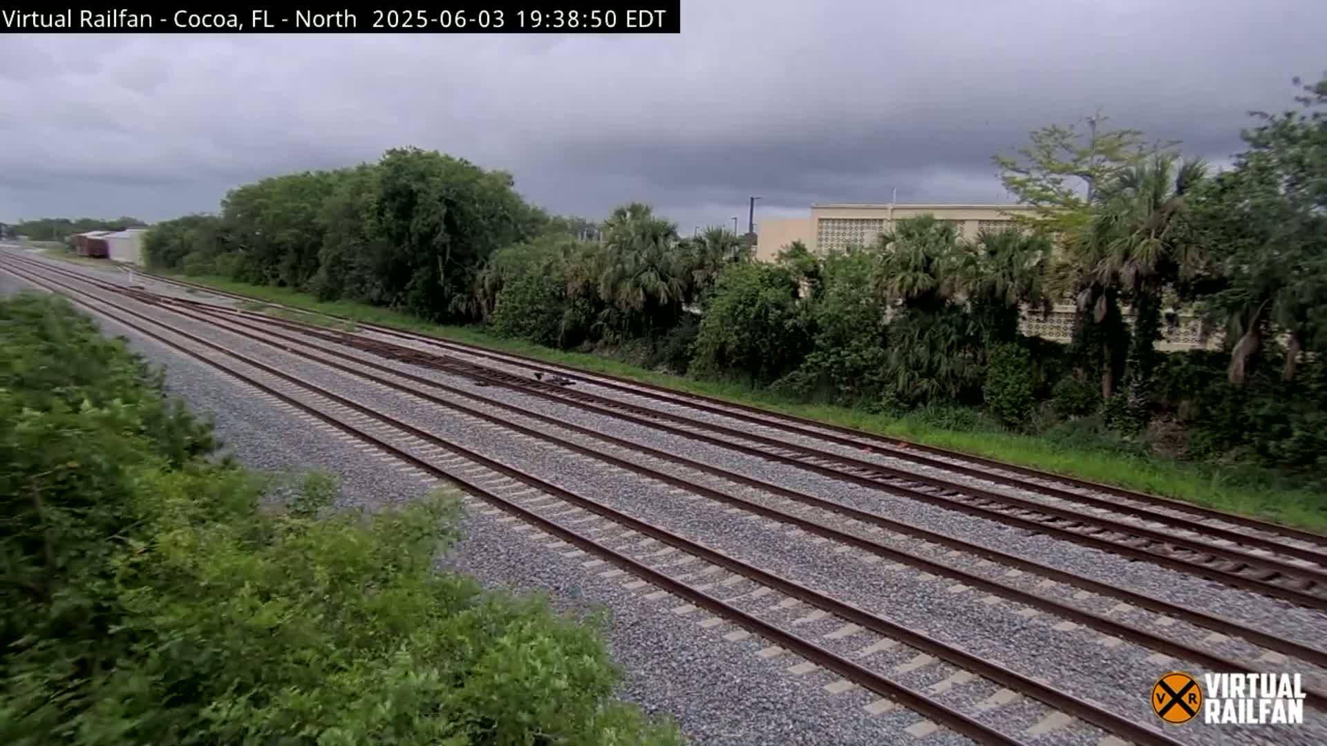 Multiple train tracks extend into the distance under an overcast sky, bordered by lush green vegetation and a building in the background.