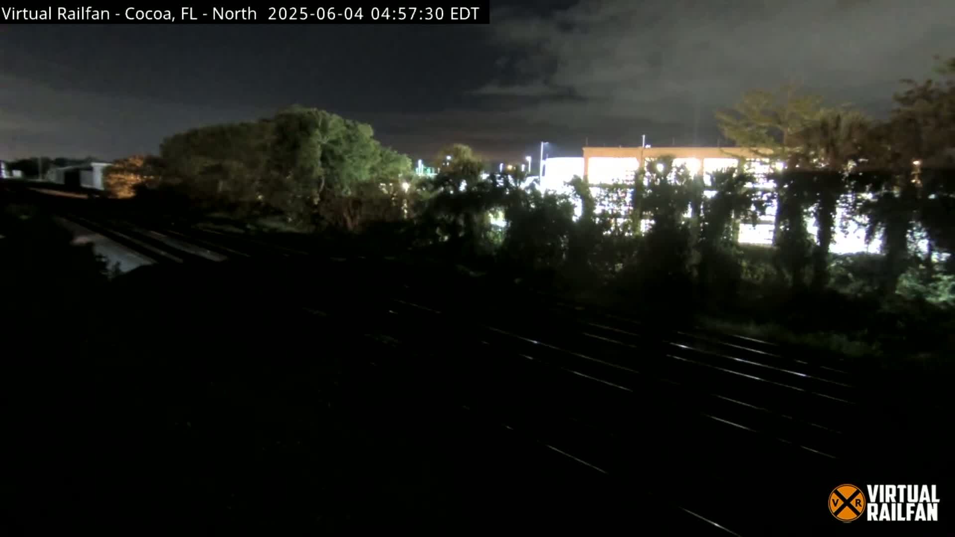 A nighttime view of train tracks next to a building partially obscured by trees under a cloudy sky.
