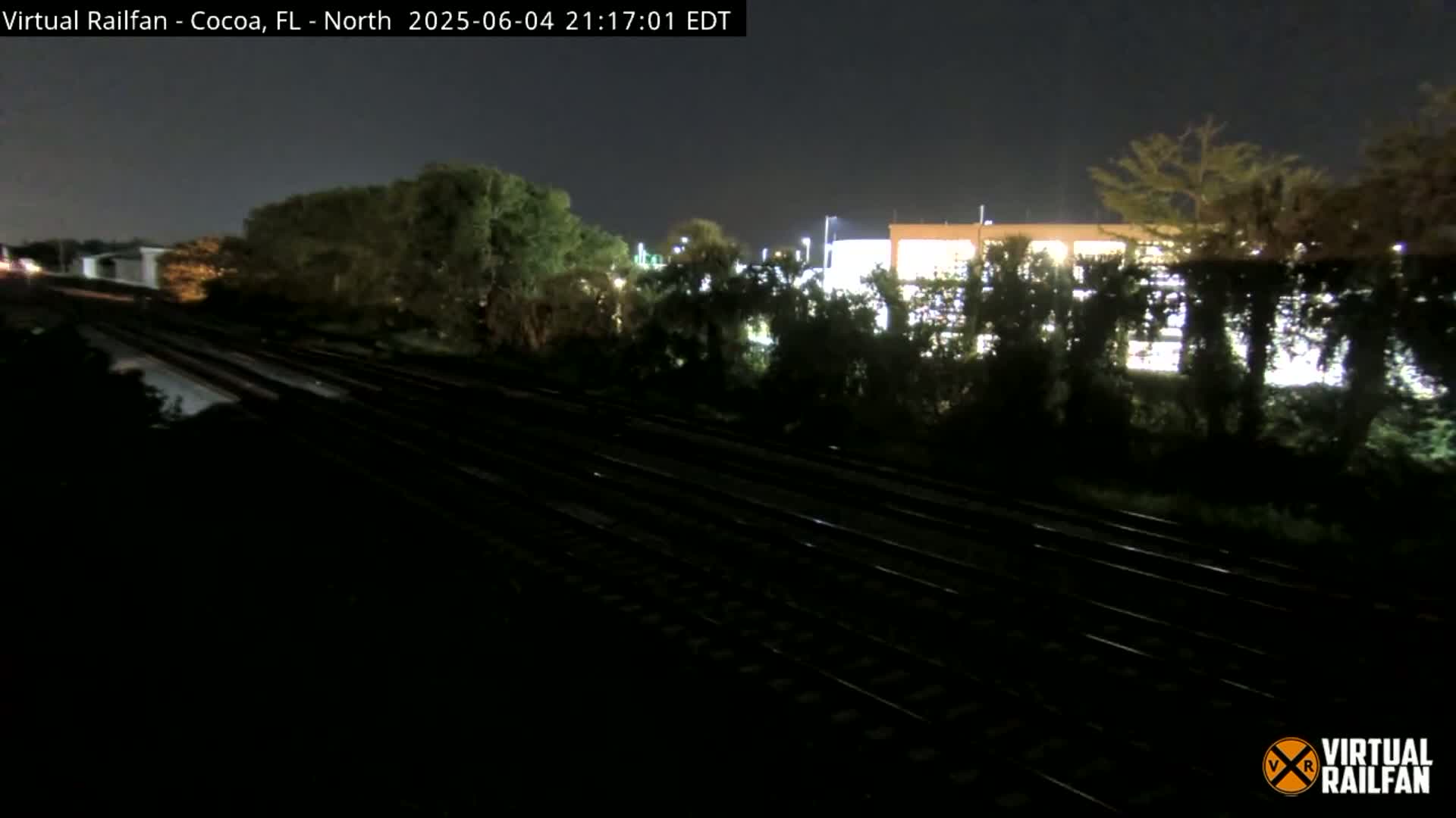 A nighttime view of train tracks running alongside a building partially obscured by trees under a dark sky.