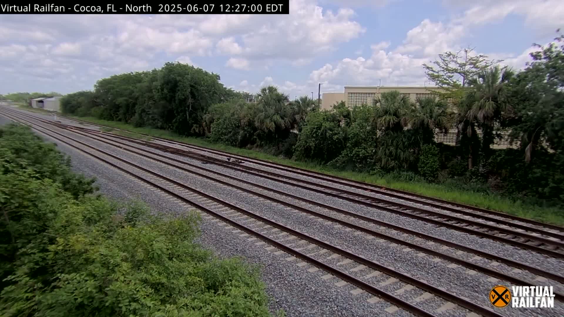Multiple train tracks stretch into the distance under a partly cloudy sky, bordered by lush green vegetation and a building in the background.