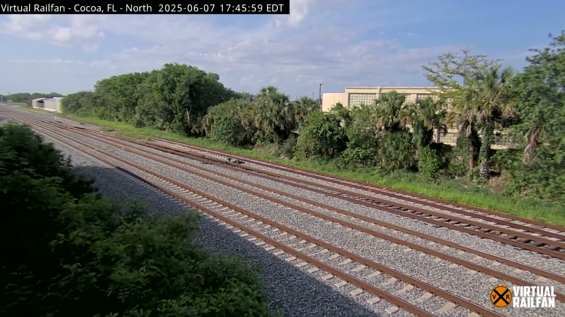 Multiple train tracks stretch into the distance on a sunny day, bordered by lush green vegetation and a building in the background.