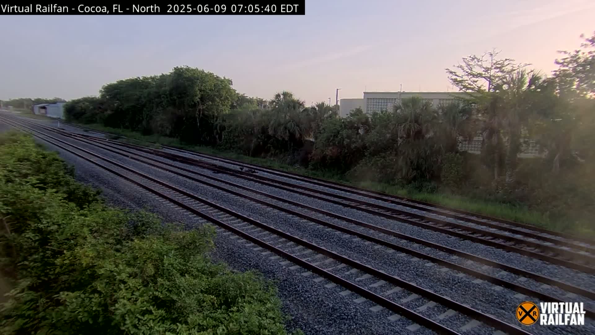 Multiple train tracks extend into the distance, bordered by lush green vegetation under a clear, dawn sky.