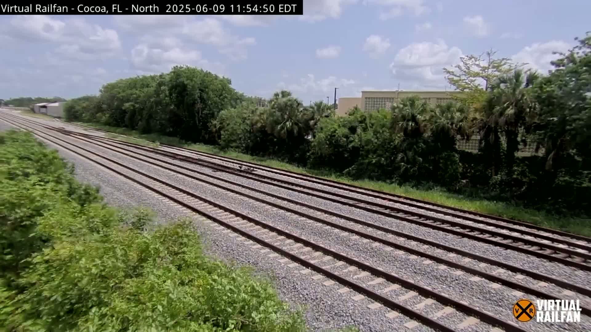Multiple parallel train tracks extend into the distance, bordered by lush green vegetation under a partly cloudy sky.