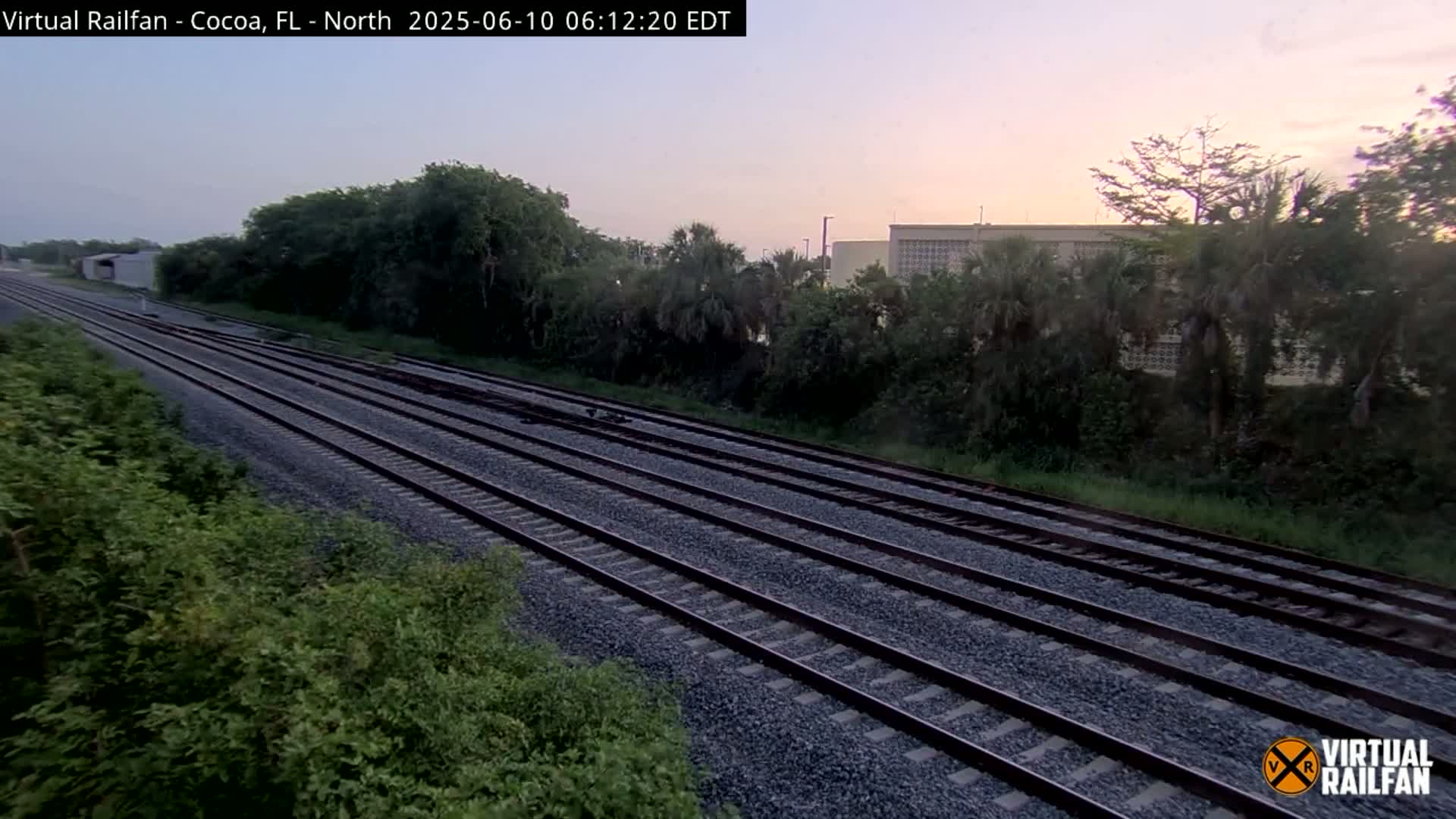 Multiple parallel train tracks extend into the distance under a dusky, pre-dawn sky, bordered by lush green vegetation and a building in the background.