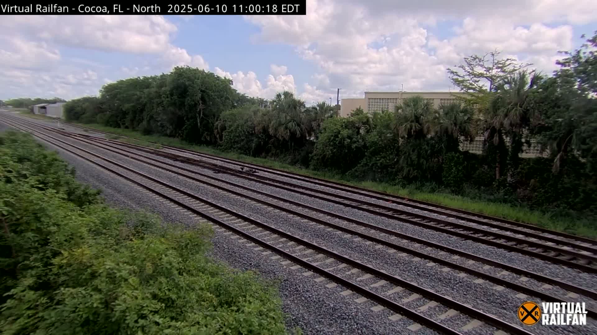 Multiple train tracks stretch into the distance under a partly cloudy sky, bordered by lush green vegetation and a building in the background.