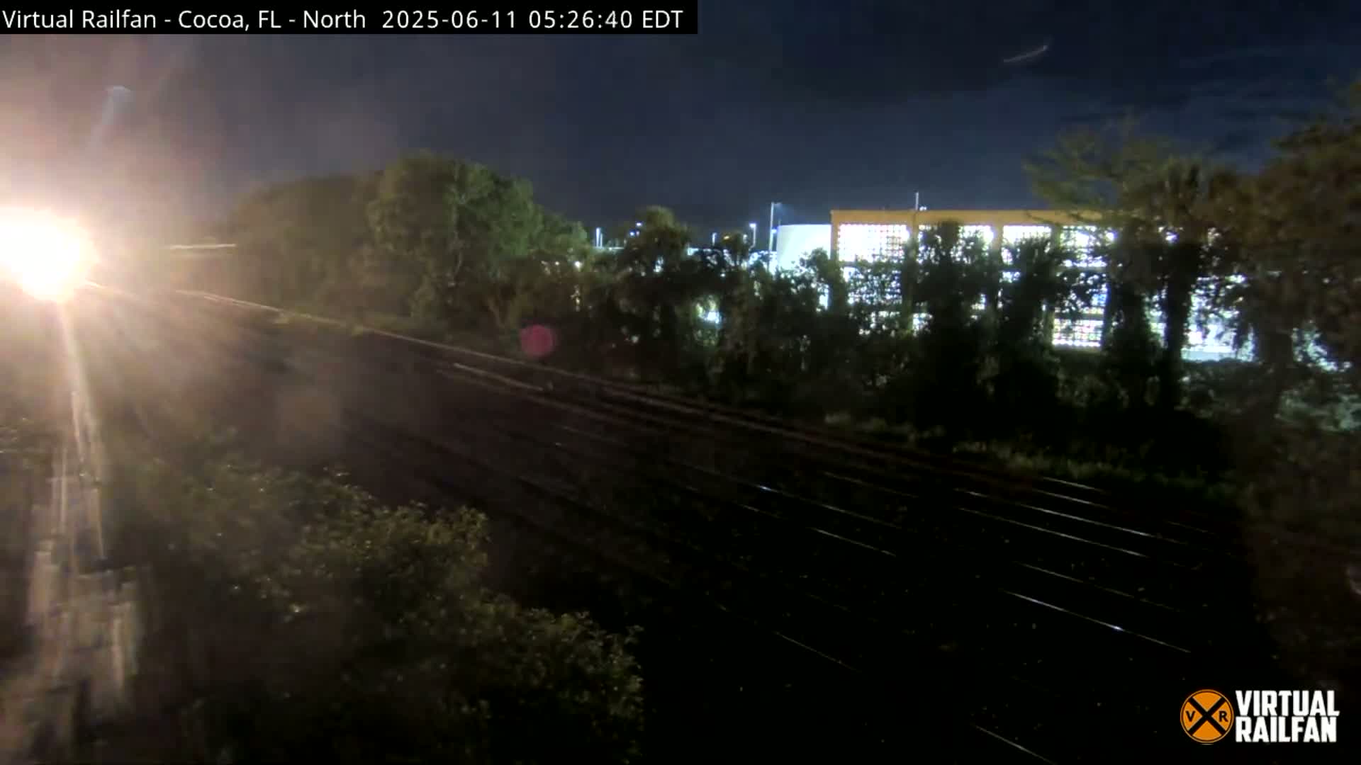 A train's headlight illuminates a nighttime scene of railroad tracks bordered by trees and a building in the distance under a dark sky.