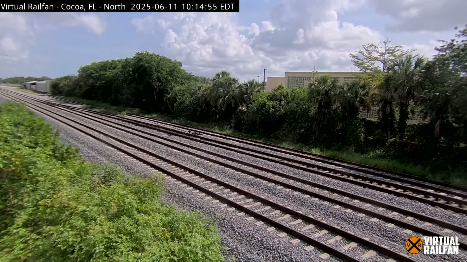 Multiple train tracks stretch into the distance under a partly cloudy sky, bordered by lush green vegetation and a building in the background.