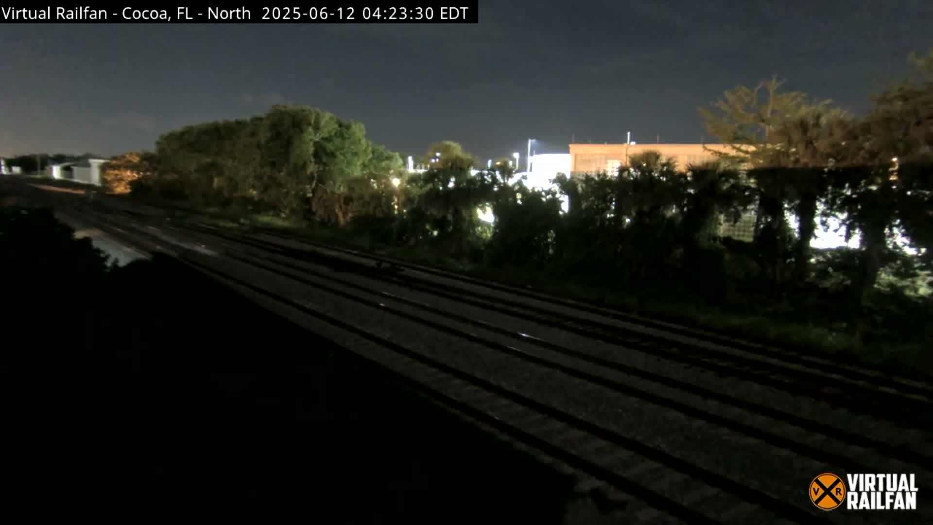 A nighttime view of multiple train tracks running alongside a line of trees and buildings under a dark sky.