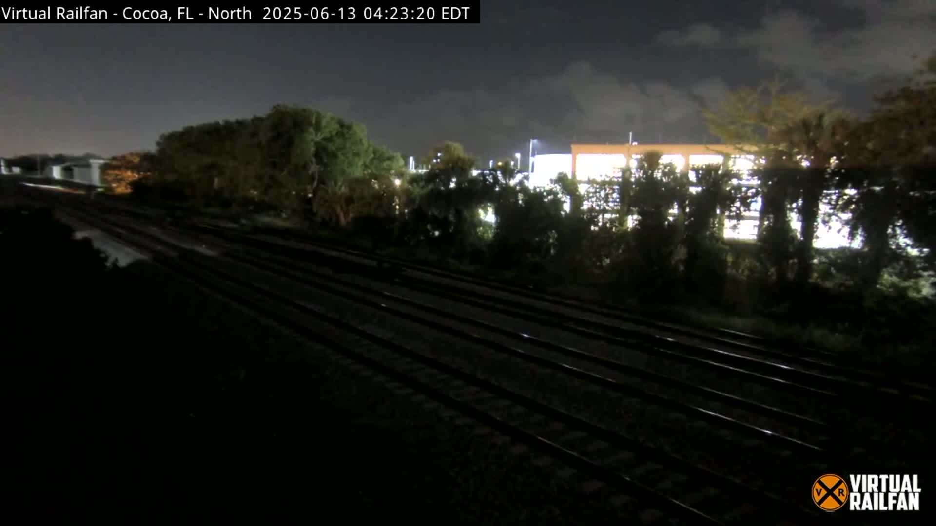 Multiple train tracks stretch into the distance under a nighttime sky, with trees and a building visible in the background.