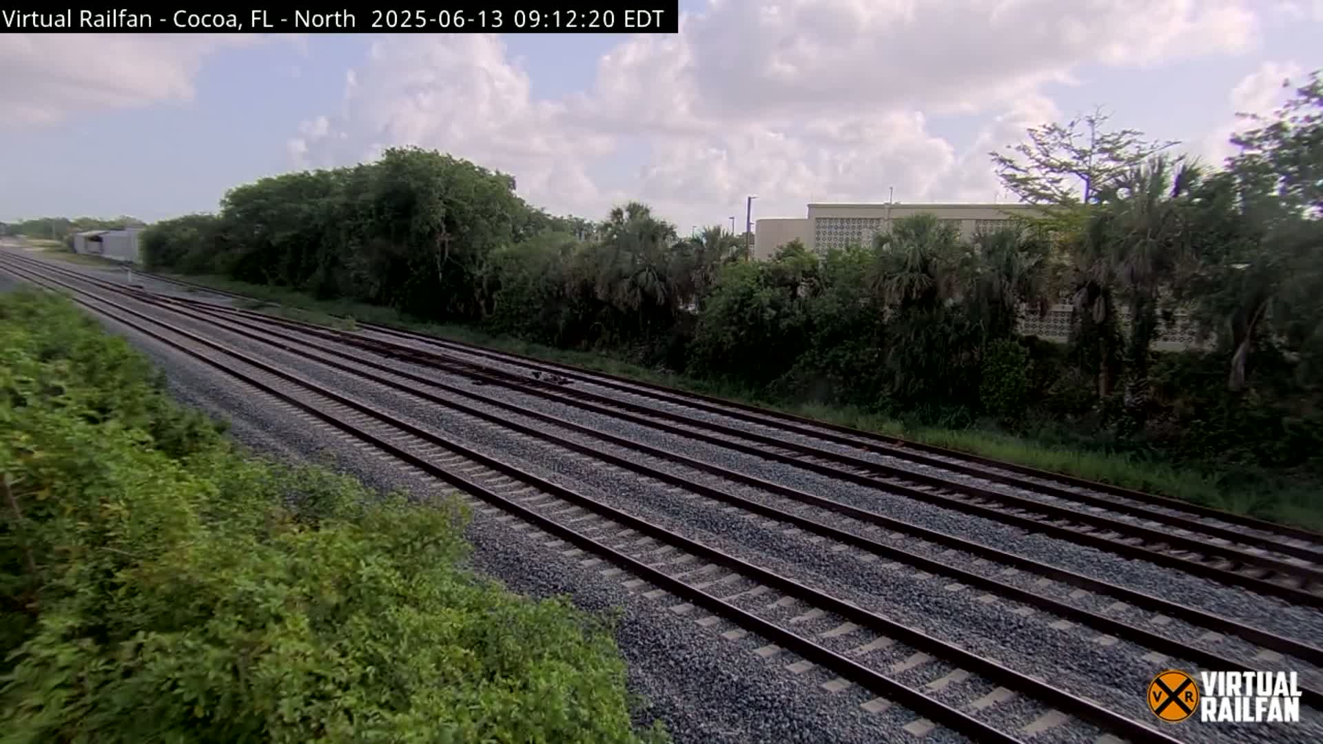 Multiple train tracks stretch into the distance, bordered by lush green vegetation under a partly cloudy sky.
