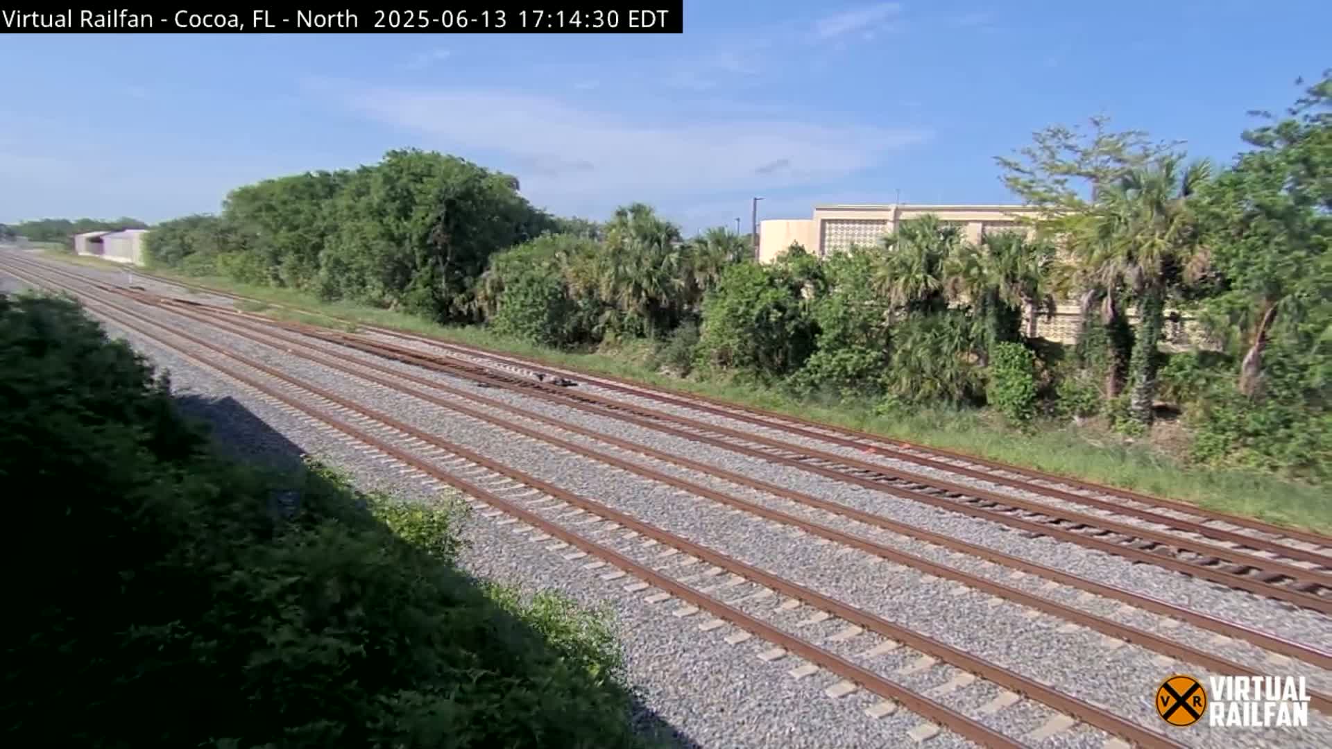 Multiple train tracks stretch into the distance under a partly sunny sky, bordered by lush green vegetation and a building in the background.