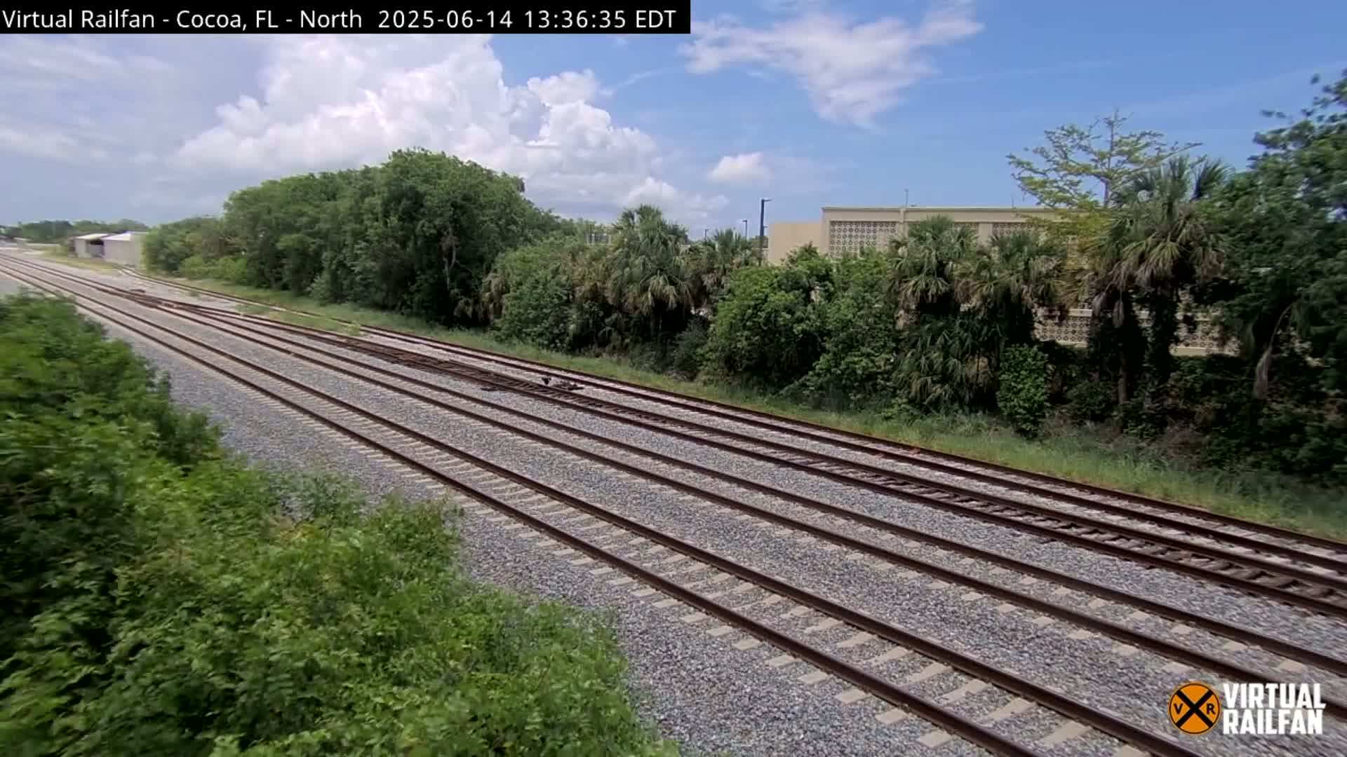 Multiple train tracks extend into the distance under a partly cloudy sky, bordered by lush green vegetation and a building.