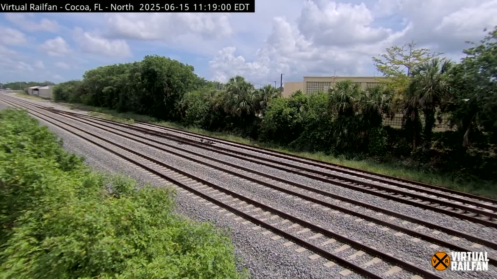 Multiple train tracks stretch into the distance under a partly cloudy sky, bordered by lush green vegetation.