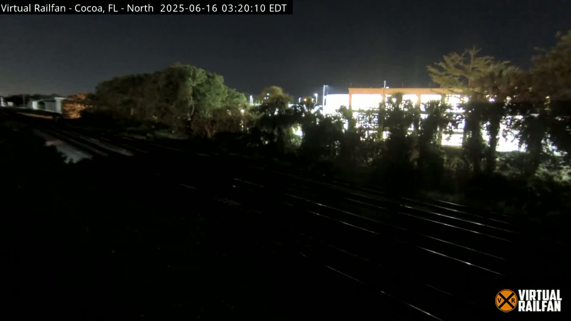 At night, a nighttime view shows train tracks leading towards a building partially obscured by trees.
