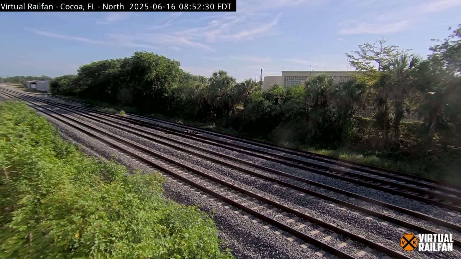 Multiple train tracks extend into the distance, bordered by lush green vegetation under a partly sunny sky.
