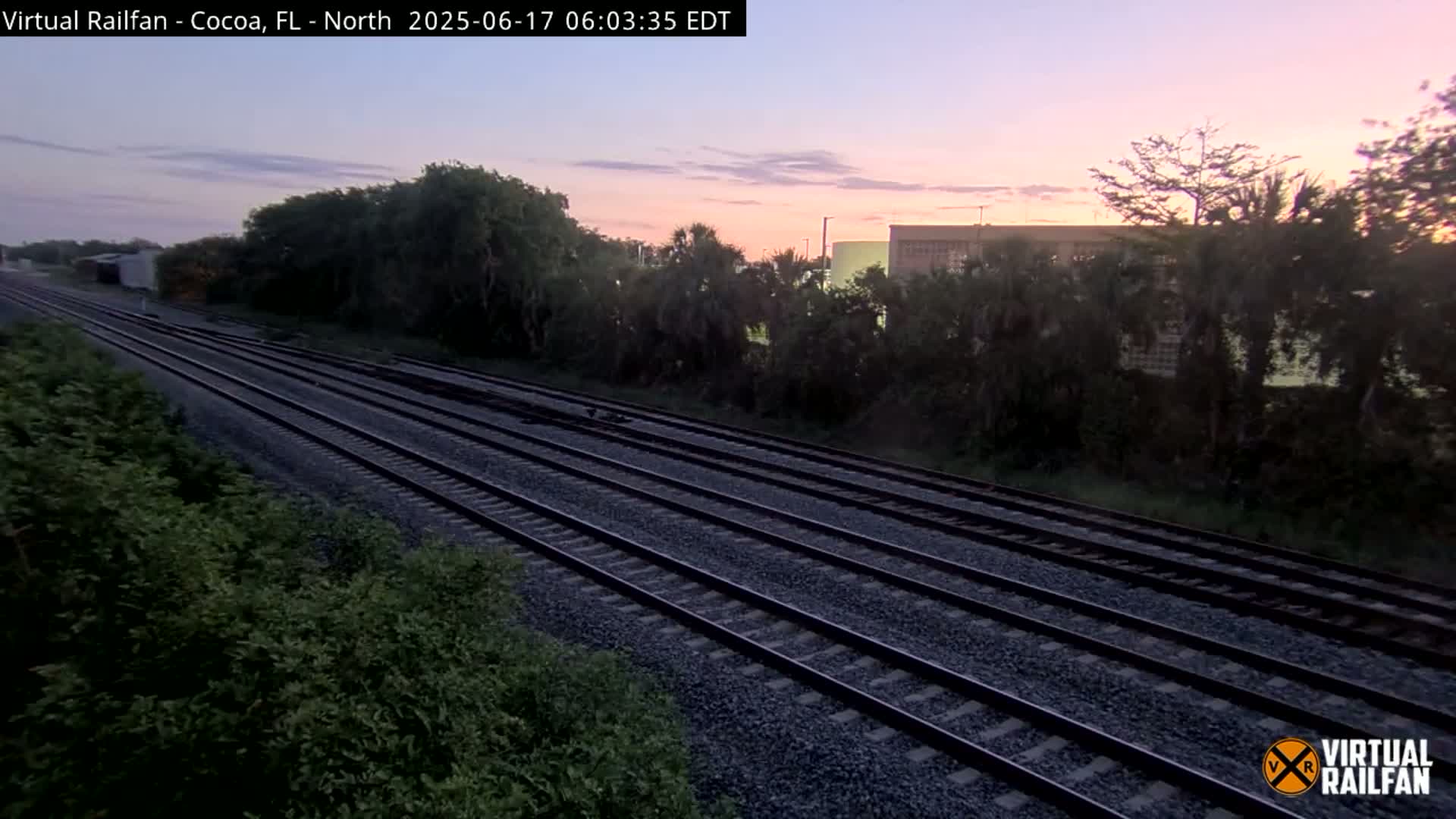 Multiple train tracks extend into the distance under a twilight sky, bordered by lush green vegetation.