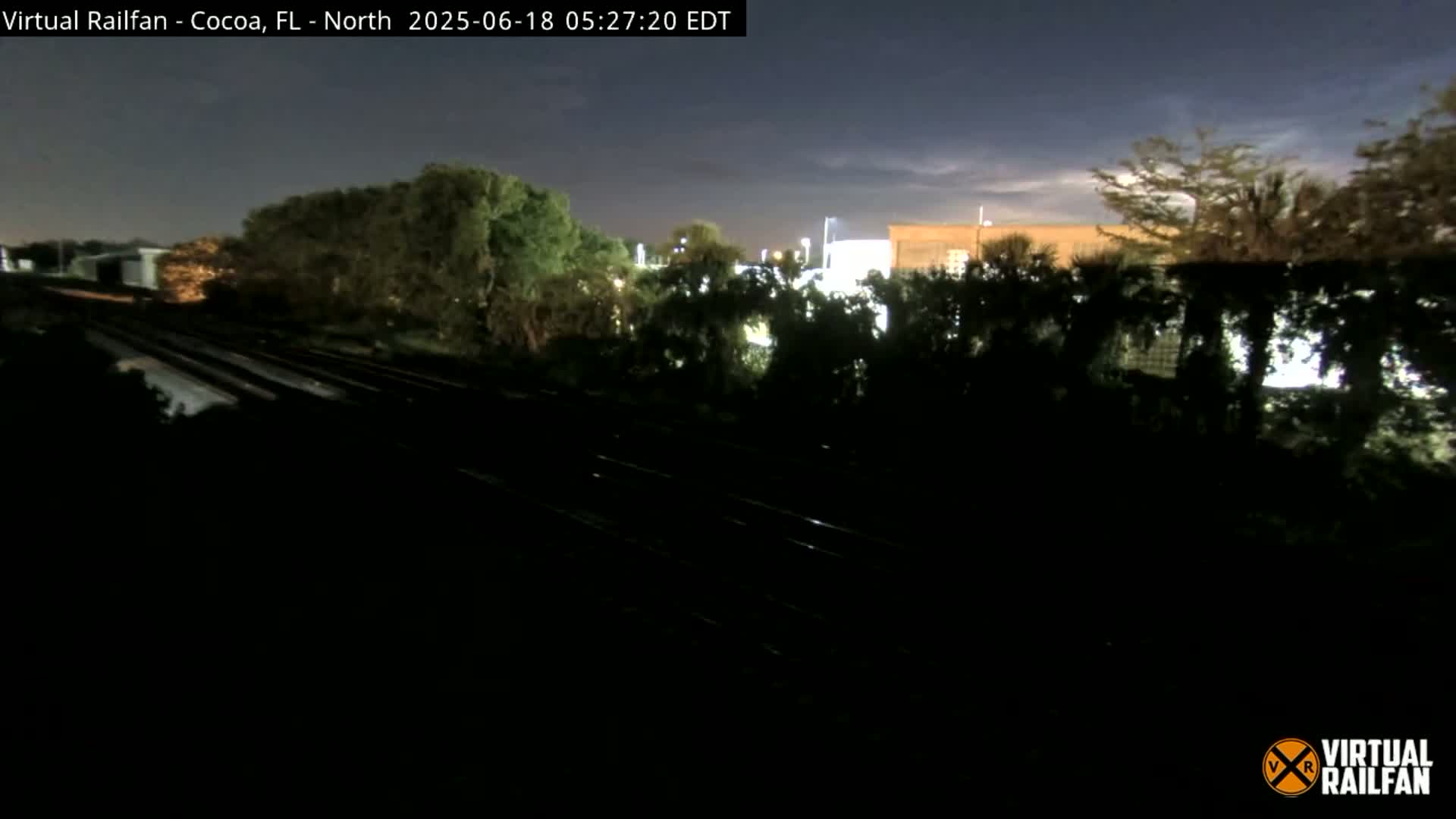 A nighttime view of railroad tracks running through a landscape of dark trees and a dimly lit building in the distance under a mostly clear sky.