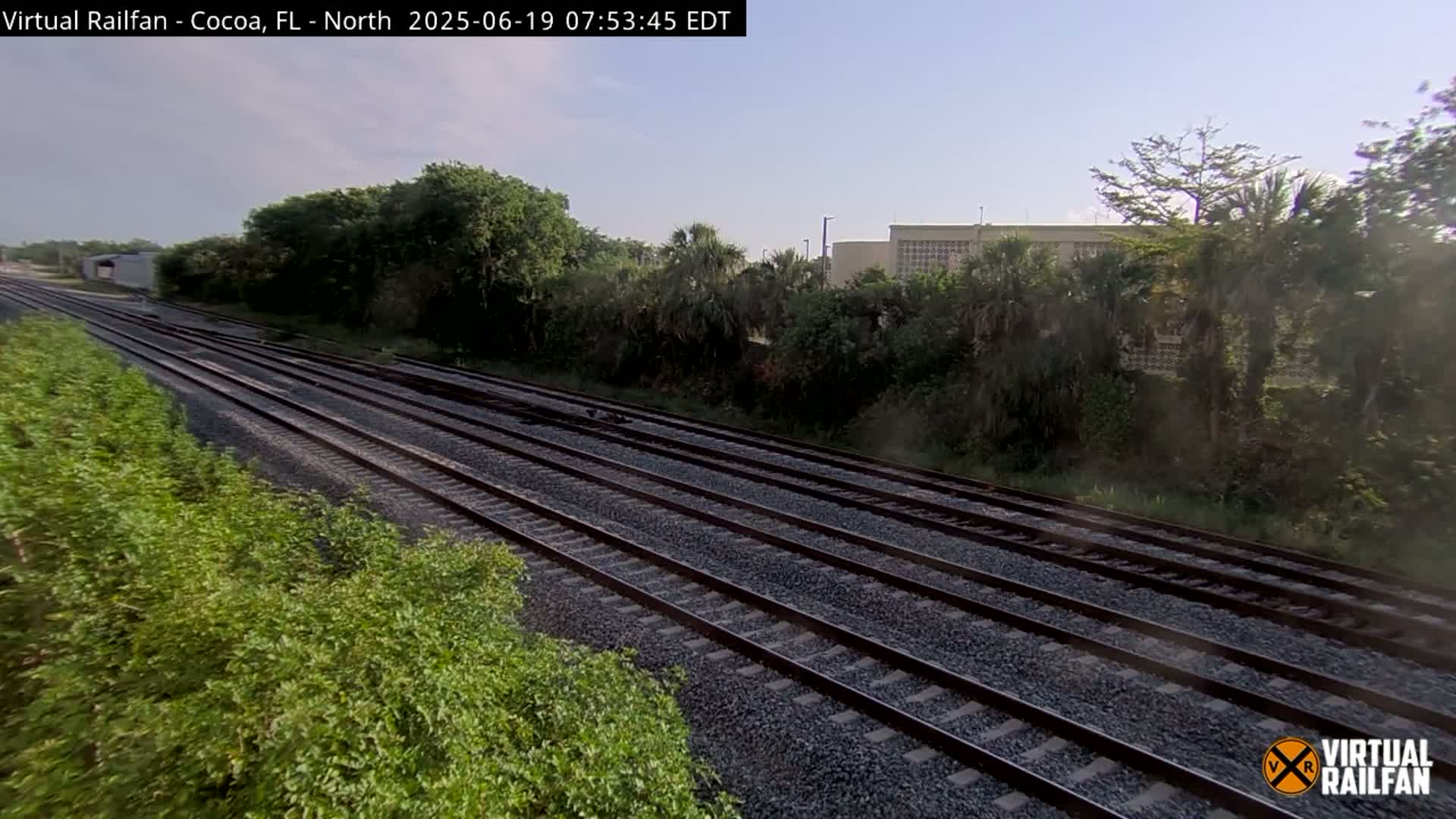 Multiple train tracks extend into the distance, bordered by lush green vegetation under a clear, sunny sky.
