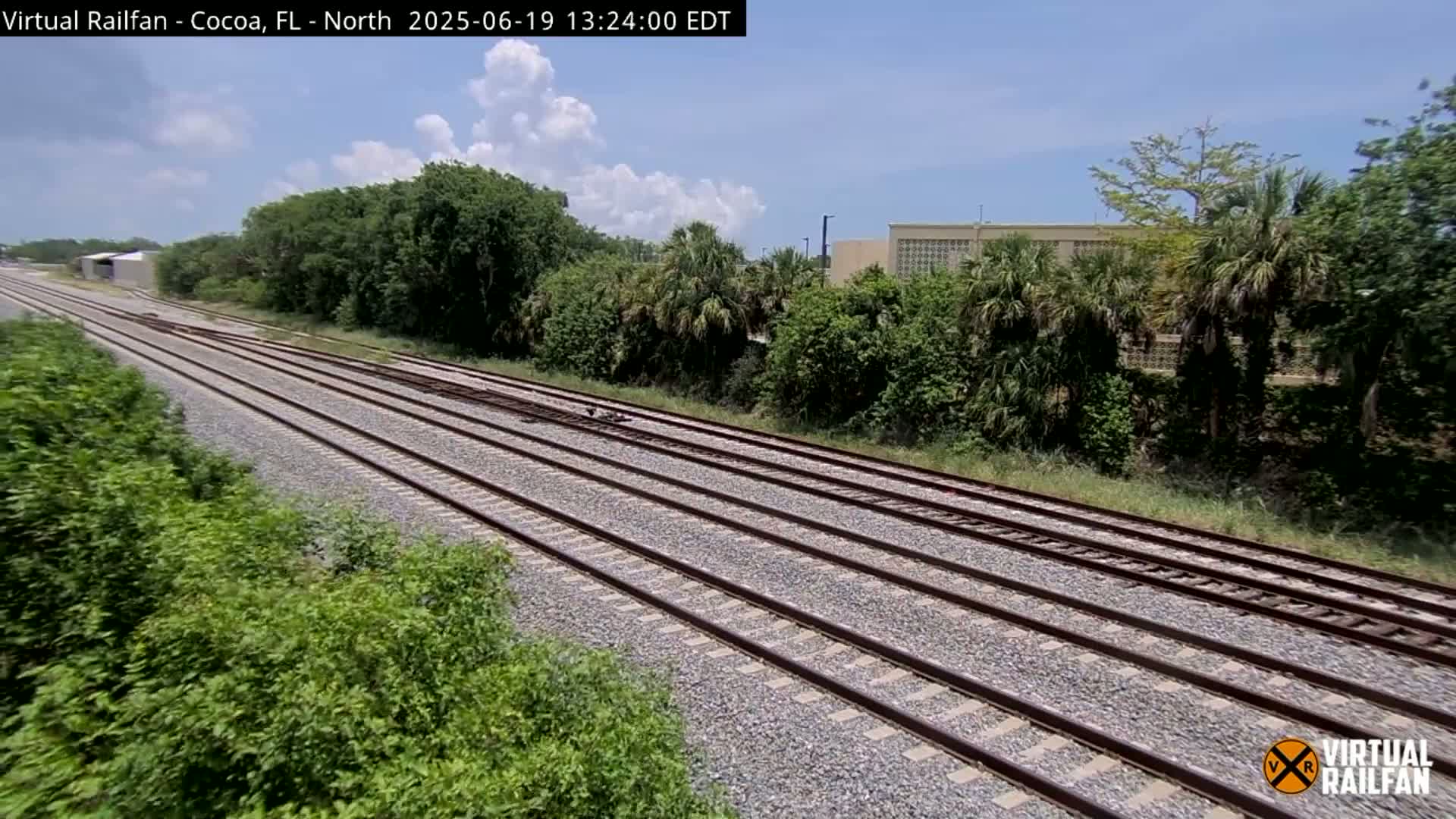 Multiple train tracks extend into the distance, bordered by lush green vegetation under a partly cloudy sky.