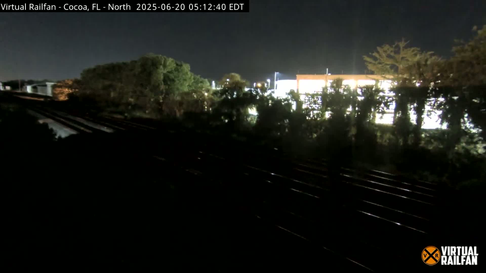 A nighttime view of train tracks running alongside a building partially obscured by trees under a clear sky.