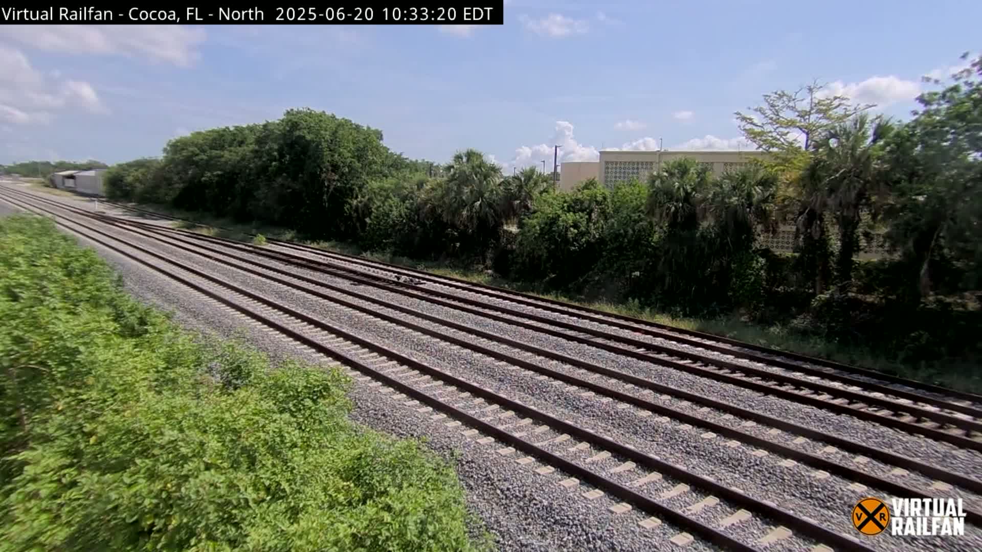 Multiple train tracks stretch into the distance under a partly cloudy sky, bordered by lush green vegetation and a building in the background.