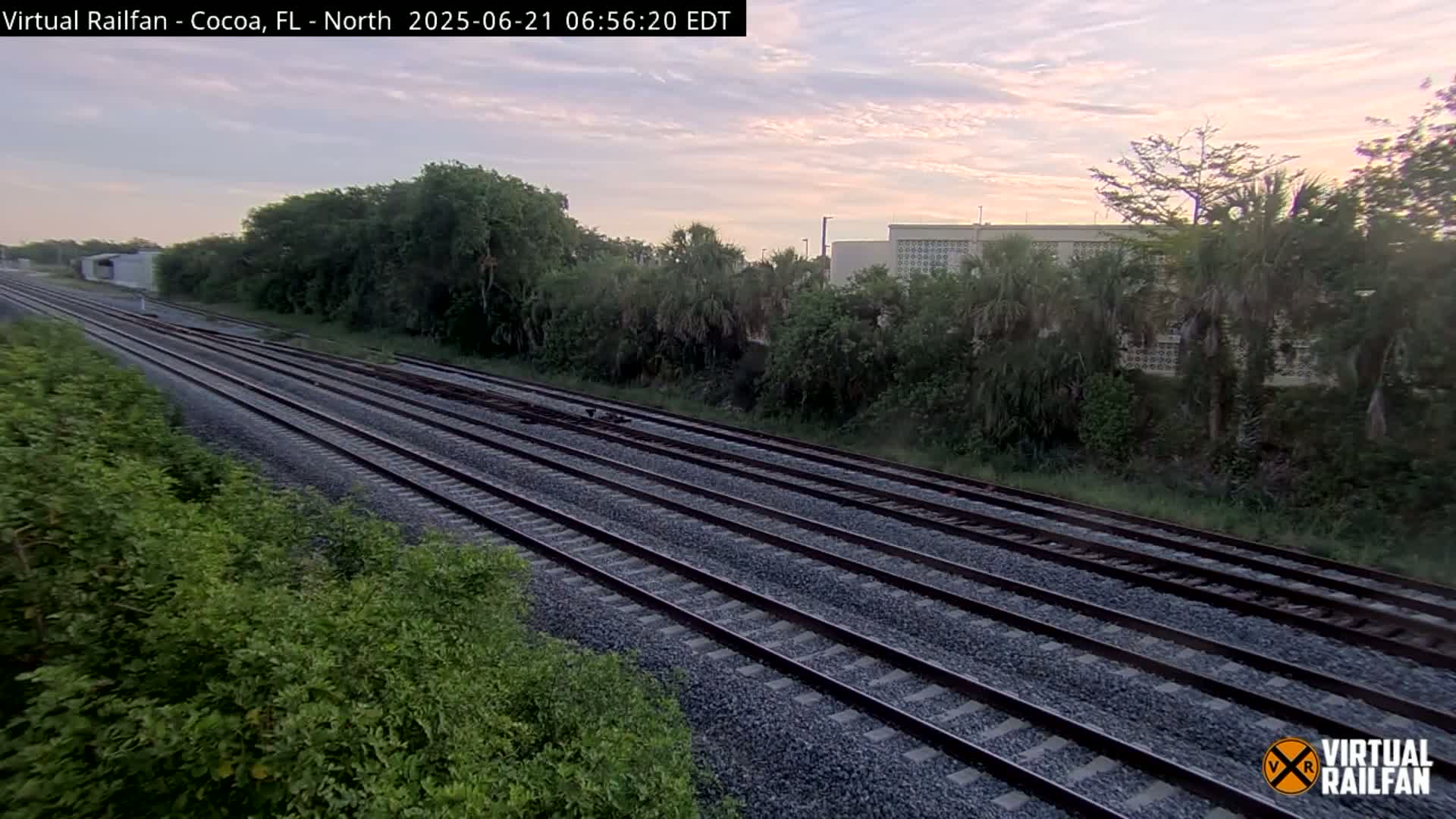 Multiple train tracks stretch into the distance, bordered by lush green vegetation under a pale pink and gray sunrise sky.
