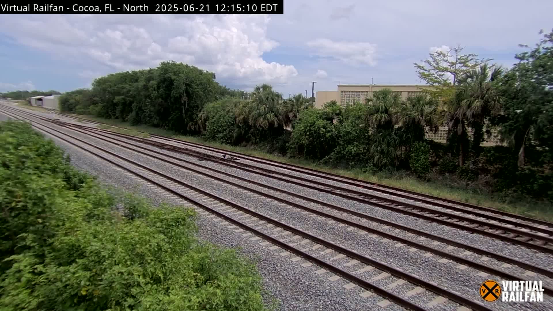 Multiple train tracks extend into the distance, bordered by lush green vegetation under a partly cloudy sky.
