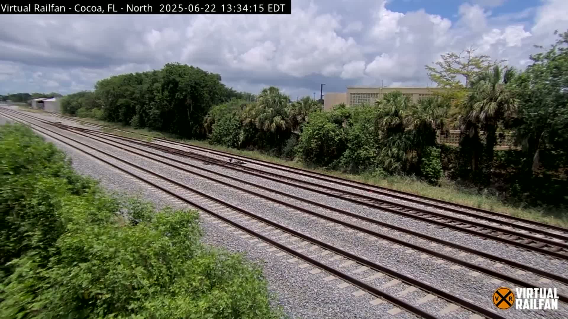 Multiple train tracks stretch into the distance, bordered by lush green vegetation under a partly cloudy sky.