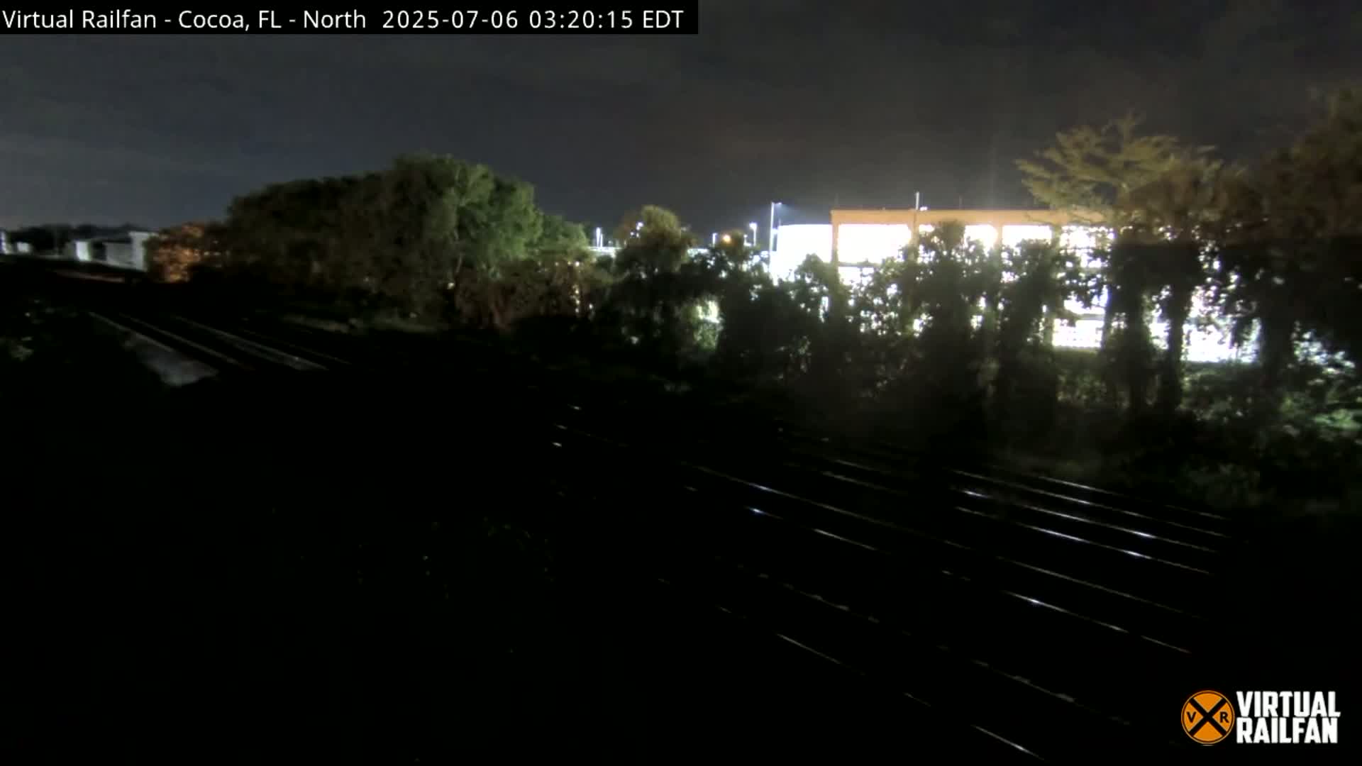A nighttime view of railroad tracks running alongside a line of trees and a brightly lit building.