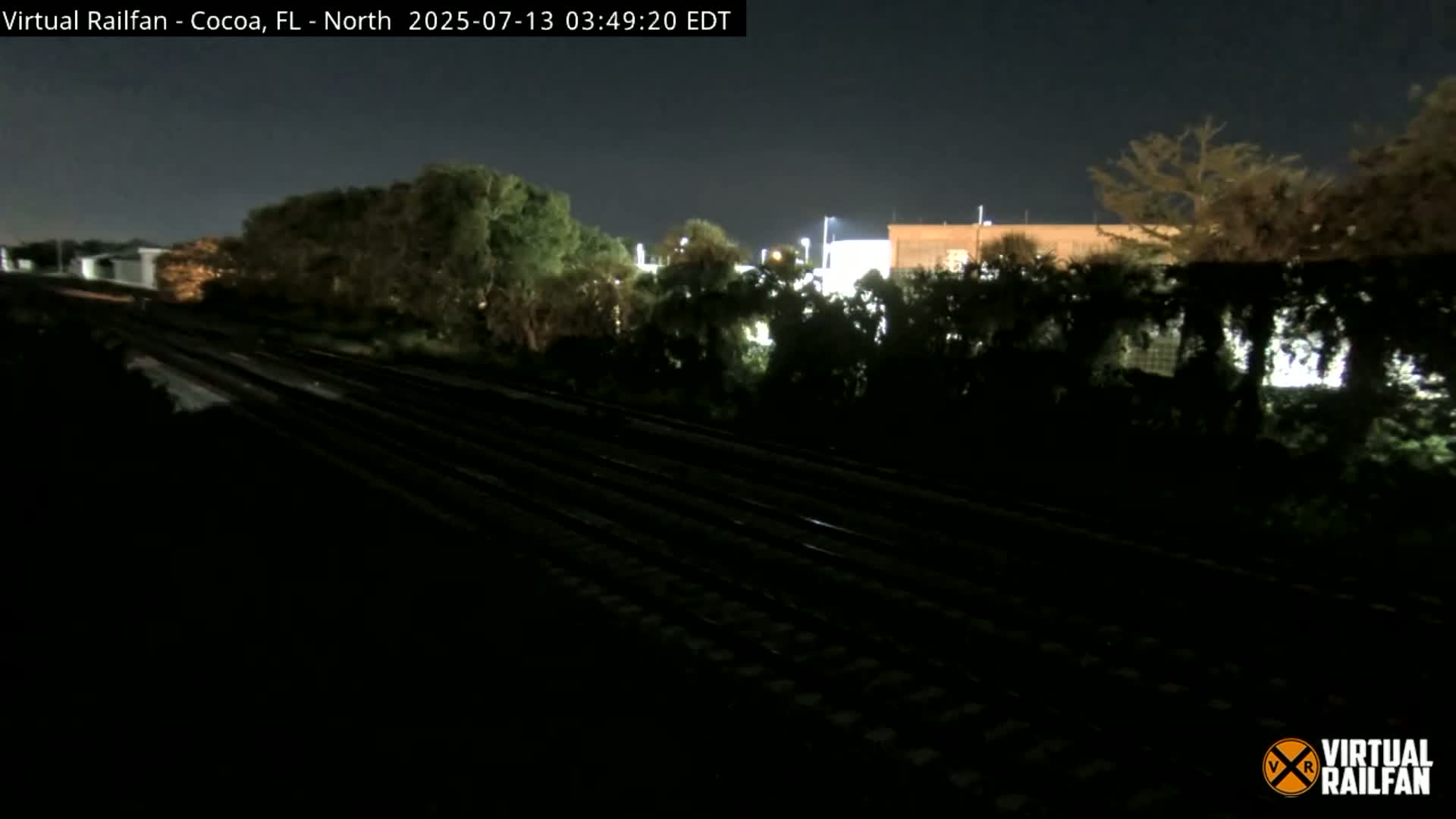 A nighttime view of railroad tracks running alongside dark vegetation and a dimly lit building under a clear sky.