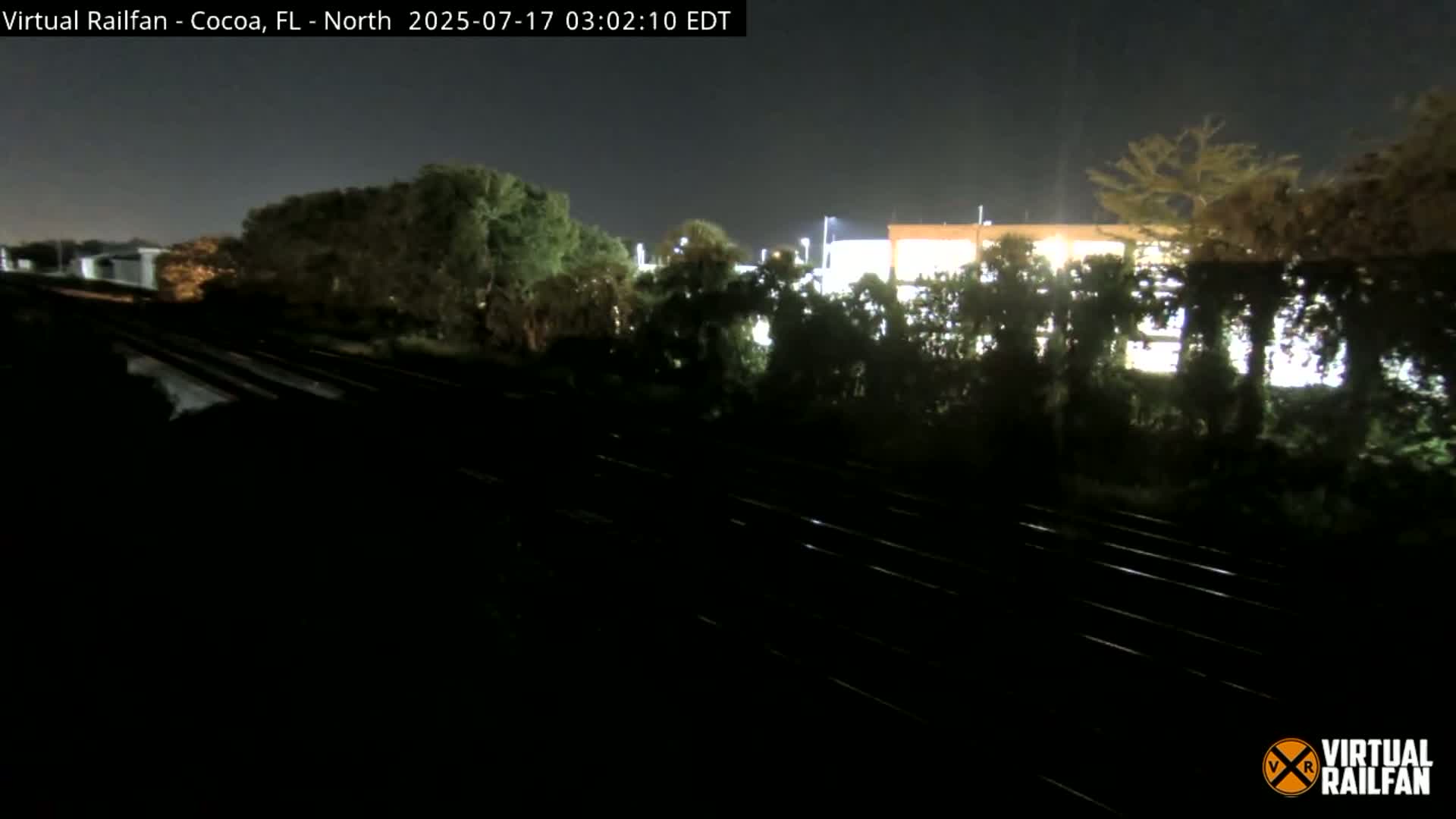 At night, train tracks run past a building partially obscured by trees under a dark sky.