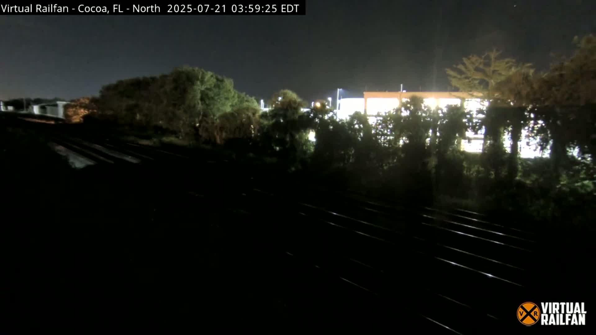 A nighttime view of railroad tracks running alongside a building partially obscured by trees under a clear sky.